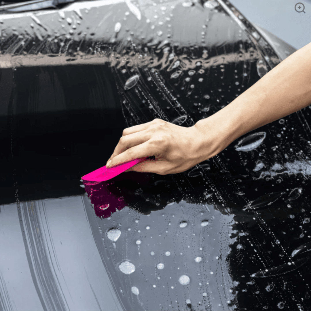 Hand using pink applicator to clean water droplets on a black car surface.